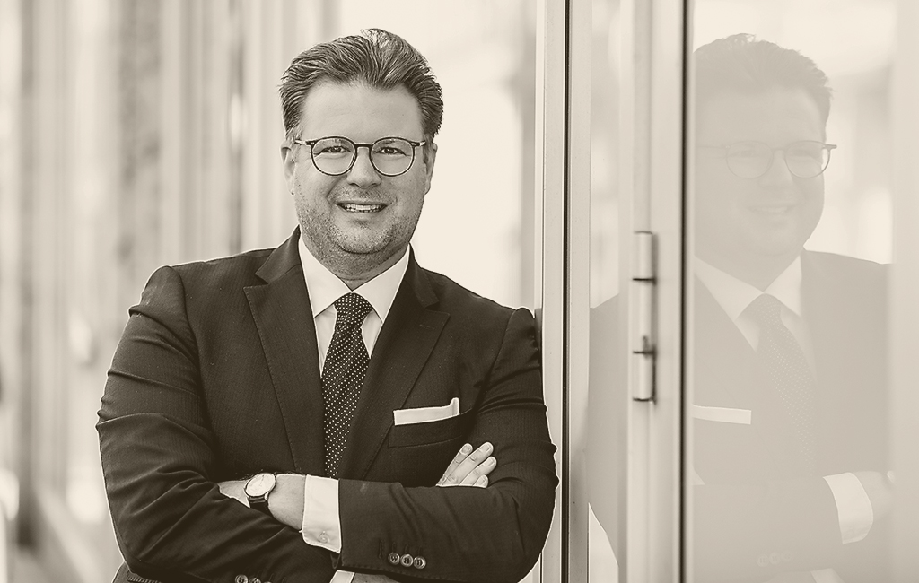 Dr. Staccioli im Anzug lächelt, lehnt an Glas, Spiegelbild sichtbar. Moderner, unscharfer Hintergrund; Schwarz-Weiß-Foto vermittelt Eleganz.